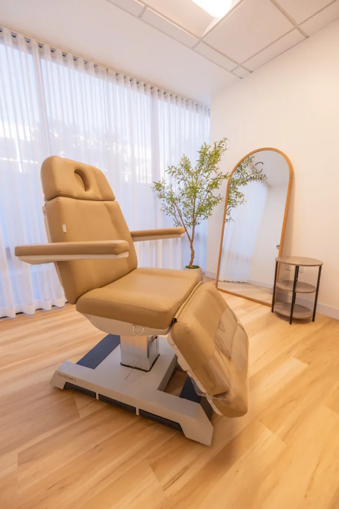 Treatment room with beige treatment chair, full-length mirror, sheer curtains, and a potted plant.
