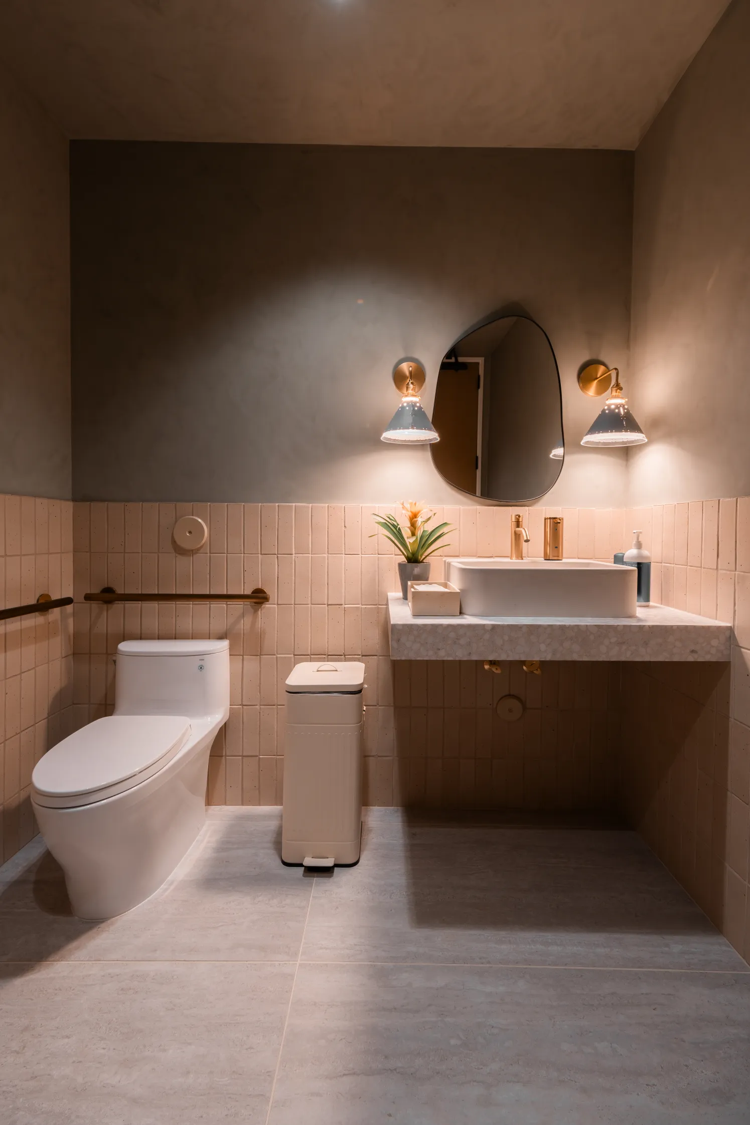 Modern restroom with neutral tones, a wall-mounted sink, oval mirror, and decorative lighting.